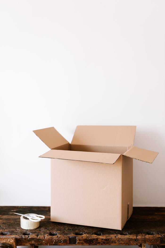 An open cardboard box on a weathered wooden table with tape and scissors beside it, ready for packing.